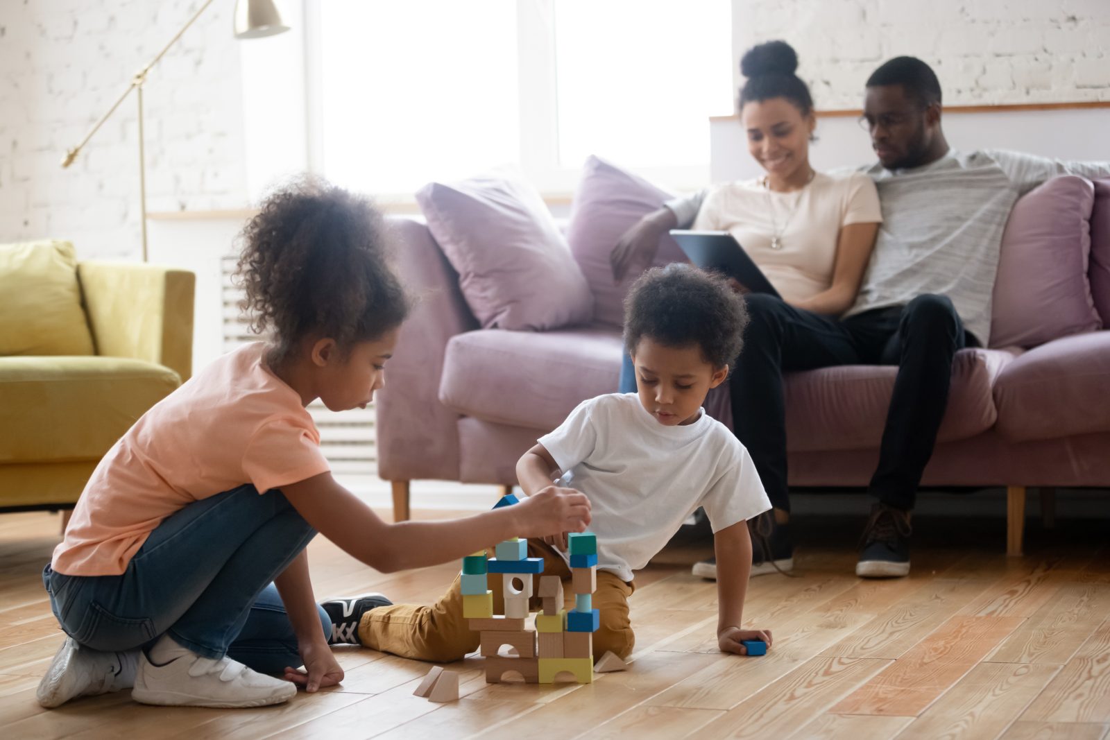 Calm bliss. Two little african kids sister and brother playing on wooden heated floor at living room building toy house from blocks and parents mum and dad sitting on sofa, hugging and using tablet pc