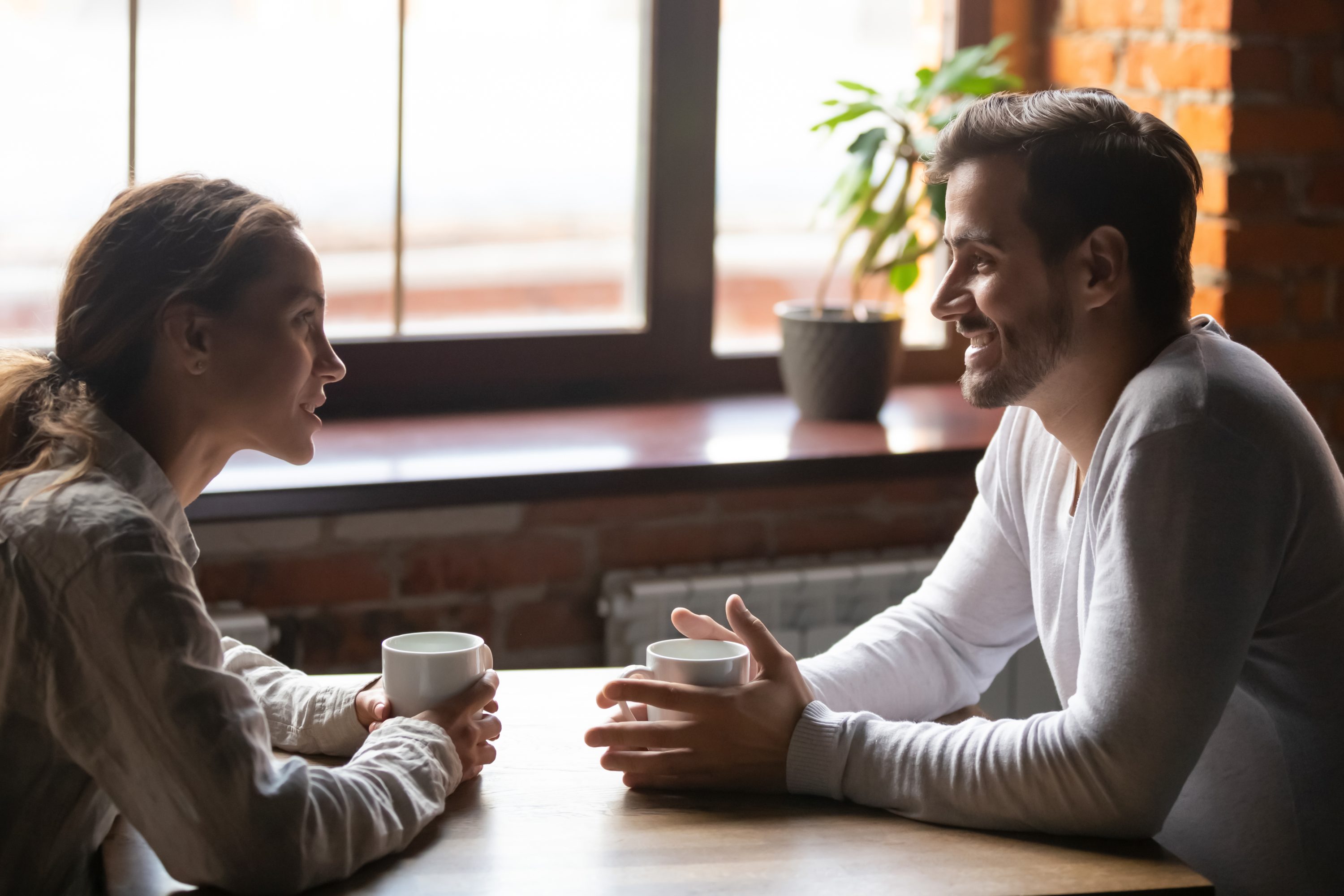 Couple sitting in cafe talking drinking tea or coffee Side view smiling biracial woman sitting at table in cafe with caucasian man couple talking in cozy coffeeshop drinking tea coffee. Heterosexual friends romantic relationships or speed dating concept